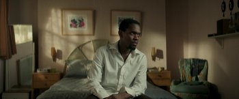 Movie still from “Yardie” (2018), directed by Idris Elba – A man sitting on top of a bed wearing a white shirt; Medium shot, High angle