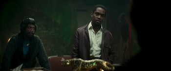 Movie still from “Yardie” (2018), directed by Idris Elba – A man standing next to a snake in a dark room; Medium shot, Low angle