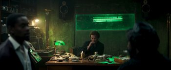 Movie still from “Yardie” (2018), directed by Idris Elba – A man sitting at a table in front of a green light; Wide shot, High angle