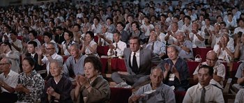 Movie still from “You Only Live Twice” (1967), directed by Lewis Gilbert – A group of people sitting in a large room clapping; Wide shot, High angle