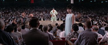 Movie still from “You Only Live Twice” (1967), directed by Lewis Gilbert – A woman in a dress standing in front of an audience; Extreme Wide shot, Over the shoulder angle
