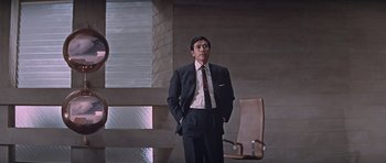 Movie still from “You Only Live Twice” (1967), directed by Lewis Gilbert – A man in a suit and tie standing next to a chair; Medium shot, Low angle