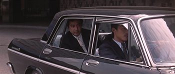 Movie still from “You Only Live Twice” (1967), directed by Lewis Gilbert – A man in a suit and tie sitting in the back seat of a car; Medium shot, Low angle