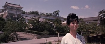 Movie still from “You Only Live Twice” (1967), directed by Lewis Gilbert – A woman standing in front of a building with trees in the background; Medium shot, Low angle