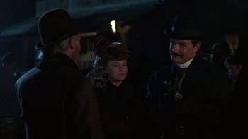 Movie still from “Young Guns” (1988), directed by Christopher Cain – Two men and a woman wearing hats in a dimly - lit room; Medium shot, Over the shoulder angle