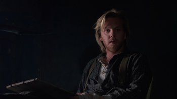 Movie still from “Young Guns” (1988), directed by Christopher Cain – A man with long blonde hair sitting in a dark room; Medium shot, Over the shoulder angle