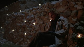Movie still from “Young Guns” (1988), directed by Christopher Cain – A person sitting on a rock wall with candles; Wide shot, High angle