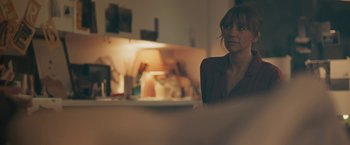 Movie still from “Zoe” (2018), directed by Drake Doremus – A woman sitting at a table in front of a lamp; Medium shot, Over the shoulder angle
