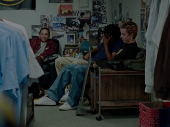 Movie still from “Mid90s” (2018), directed by Jonah Hill – A group of people sitting in a room with a skateboard; Wide shot, High angle