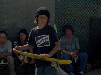 Movie still from “Mid90s” (2018), directed by Jonah Hill – A young boy holding a yellow skateboard in front of two other people; Medium shot, Over the shoulder angle