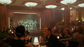 Movie still from “Bullets Over Broadway” (1994), directed by Woody Allen – A man and a woman sitting at a table in front of an audience; Wide shot, High angle