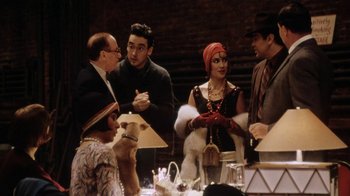 Movie still from “Bullets Over Broadway” (1994), directed by Woody Allen – A group of people standing around a table with food; Medium shot, Over the shoulder angle