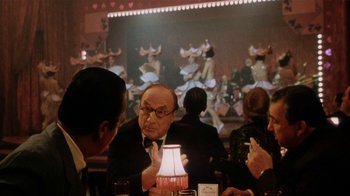 Movie still from “Bullets Over Broadway” (1994), directed by Woody Allen – An older man sitting at a table with a lamp in front of him; Medium shot, Over the shoulder angle