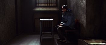 Movie still from “12” (2007), directed by Nikita Mikhalkov – A person sitting on a bench in a room; Wide shot, High angle