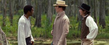 Movie still from “12 Years a Slave” (2013), directed by Steve McQueen – A man wearing a straw hat and a beige jacket standing next to another man in a field; Medium shot, Over the shoulder angle