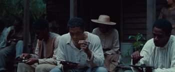 Movie still from “12 Years a Slave” (2013), directed by Steve McQueen – A man sitting on the ground while holding a cell phone; Medium shot, High angle