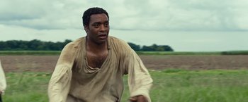 Movie still from “12 Years a Slave” (2013), directed by Steve McQueen – A black man in a brown shirt is running in a field; Medium shot, Over the shoulder angle