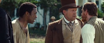 Movie still from “12 Years a Slave” (2013), directed by Steve McQueen – A man in a top hat standing next to another man in a suit; Medium shot, Over the shoulder angle