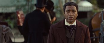 Movie still from “12 Years a Slave” (2013), directed by Steve McQueen – A man wearing a suit and a top hat in front of other men; Close Up shot, Over the shoulder angle