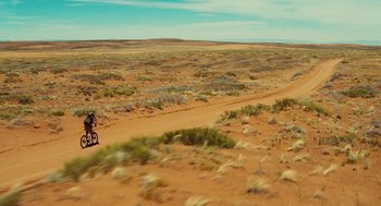Movie still from “127 Hours” (2010), directed by Danny Boyle – A person riding a bike on a dirt road near a field; Extreme Wide shot, High angle