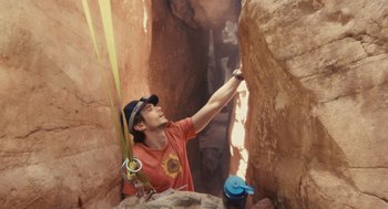Movie still from “127 Hours” (2010), directed by Danny Boyle – A man in a hat is reaching up to reach something in a cave; Medium shot, Low angle
