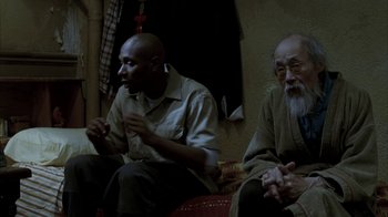 Movie still from “16 Blocks” (2006), directed by Richard Donner – Two men are sitting on a couch and one of them is talking to another man; Medium shot, Over the shoulder angle