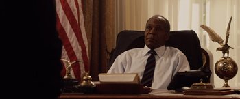 Movie still from “2012” (2009), directed by Roland Emmerich – A man sitting at a desk in front of an american flag; Medium shot, Low angle