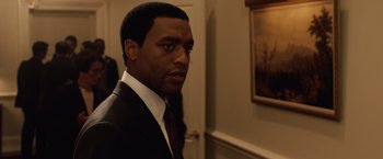 Movie still from “2012” (2009), directed by Roland Emmerich – A man in a suit standing in front of a wall; Medium shot, Over the shoulder angle