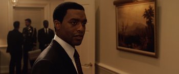 Movie still from “2012” (2009), directed by Roland Emmerich – A man wearing a suit and tie standing in front of a painting; Medium shot, Over the shoulder angle