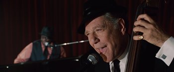 Movie still from “2012” (2009), directed by Roland Emmerich – An older man wearing a black hat and a microphone; Close Up shot, Over the shoulder angle