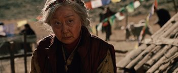 Movie still from “2012” (2009), directed by Roland Emmerich – An older woman wearing a headdress and a red jacket; Close Up shot, Over the shoulder angle