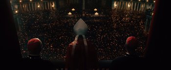 Movie still from “2012” (2009), directed by Roland Emmerich – A person wearing a priest's outfit and a hat is standing in front of an audience; Extreme Wide shot, High angle