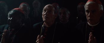 Movie still from “2012” (2009), directed by Roland Emmerich – A man in a priest's outfit praying in front of a group of men; Medium shot, Low angle