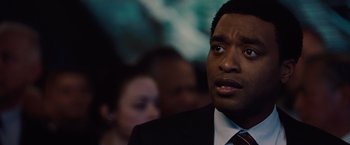 Movie still from “2012” (2009), directed by Roland Emmerich – A man in a suit and tie looking to his left; Close Up shot, Over the shoulder angle