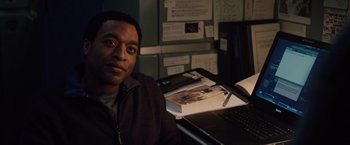 Movie still from “2012” (2009), directed by Roland Emmerich – A man sitting in front of a laptop computer on top of a desk; Medium shot, Over the shoulder angle