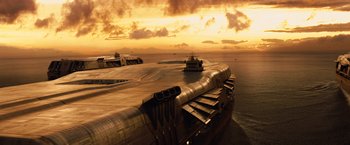 Movie still from “2012” (2009), directed by Roland Emmerich – An aerial view of an aircraft carrier at sunset; Extreme Wide shot, High angle