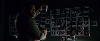 Movie still from “21” (2008), directed by Robert Luketic – A man holding a roll of paper in front of a wall of pictures; Medium shot, High angle