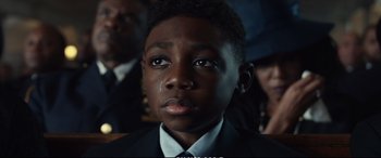 Movie still from “21 Bridges” (2019), directed by Brian Kirk – A young boy in a suit and tie looking into the camera; Close Up shot, Over the shoulder angle