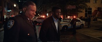 Movie still from “21 Bridges” (2019), directed by Brian Kirk – A man in a suit standing next to another man in front of a cop car; Medium shot, Over the shoulder angle