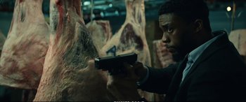 Movie still from “21 Bridges” (2019), directed by Brian Kirk – A man holding a gun in front of a bunch of meat; Medium shot, Over the shoulder angle
