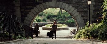 Movie still from “25th Hour” (2002), directed by Spike Lee – A man walking down a sidewalk with a suitcase under an archway; Wide shot, Over the shoulder angle