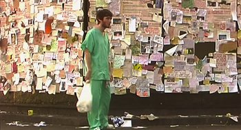 Movie still from “28 Days Later” (2002), directed by Danny Boyle – A man in green scrubs stands in front of a wall covered in papers; Wide shot, High angle