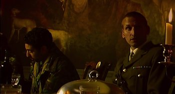 Movie still from “28 Days Later” (2002), directed by Danny Boyle – Two men sitting at a table in front of a painting; Medium shot, Low angle