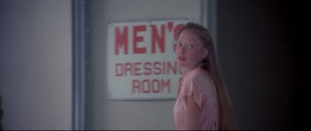 Movie still from “3 Women” (1977), directed by Robert Altman – A woman standing in front of a men's dressing room sign; Medium shot, Over the shoulder angle