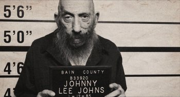 Movie still from “3 from Hell” (2019), directed by Rob Zombie – An old man with a beard is holding up a mugshot; Medium shot, Low angle