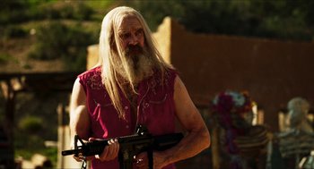 Movie still from “3 from Hell” (2019), directed by Rob Zombie – A man with long white hair and a long beard holding a machine gun; Medium shot, Over the shoulder angle
