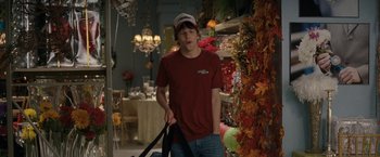 Movie still from “30 Minutes or Less” (2011), directed by Ruben Fleischer – A man in a red t - shirt holding a black bag in a room; Medium shot, Over the shoulder angle