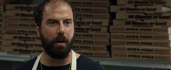 Movie still from “30 Minutes or Less” (2011), directed by Ruben Fleischer – A man with a beard wearing an apron in front of stacks of pizza boxes; Close Up shot, Over the shoulder angle