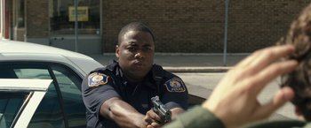 Movie still from “30 Minutes or Less” (2011), directed by Ruben Fleischer – A police officer holding a gun while sitting on a bench; Medium shot, Over the shoulder angle