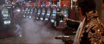 Movie still from “3000 Miles to Graceland” (2001), directed by Demian Lichtenstein – A person holding a gun in front of a row of slot machines; Wide shot, Over the shoulder angle